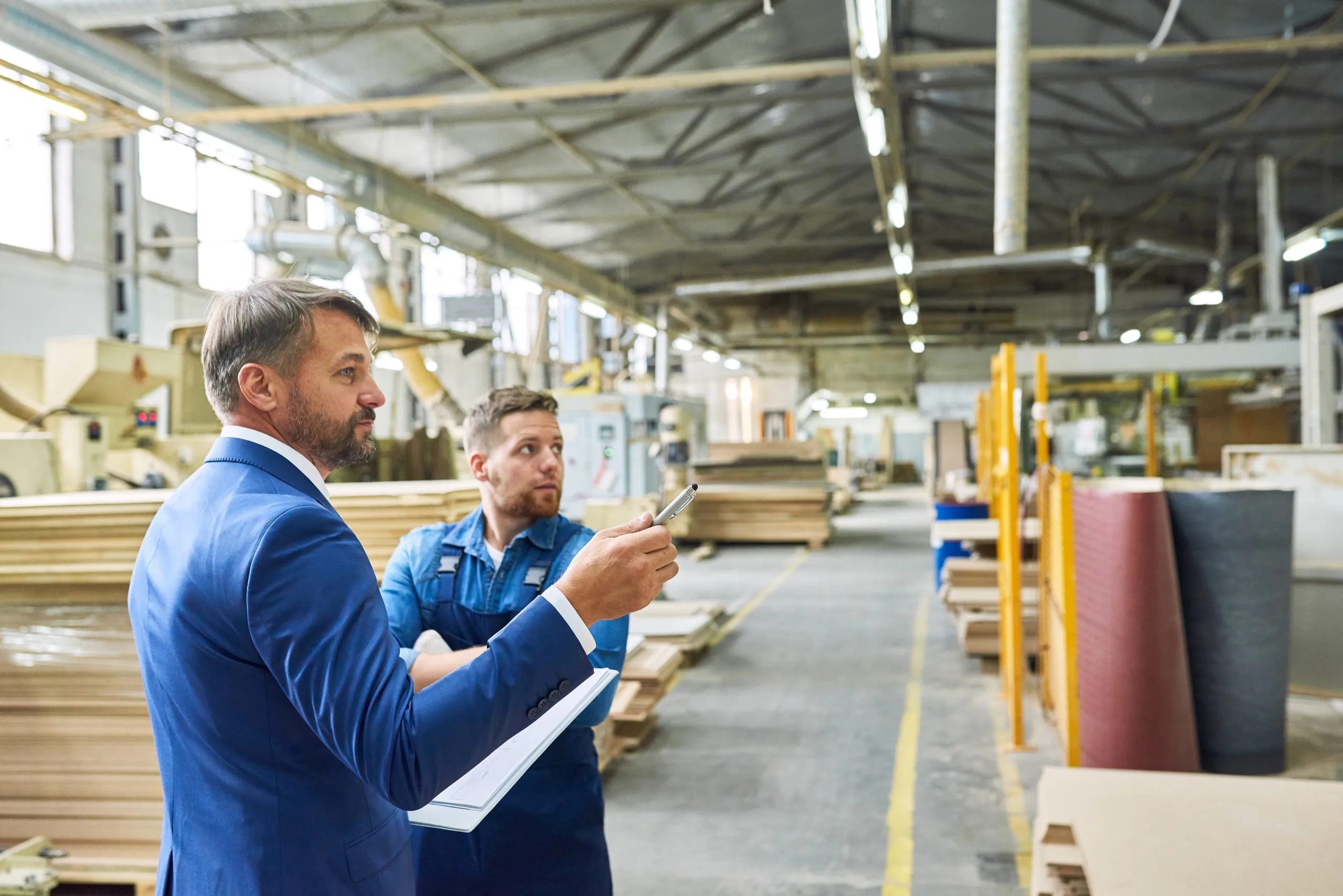 Man in a blue suit with a notepad and a worker in denim overalls inspect a woodworking factory