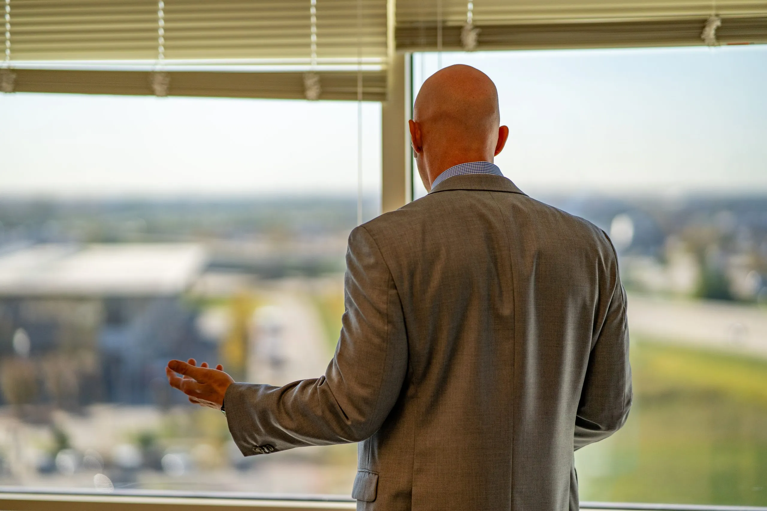 Person in a suit stands with back turned, gesturing while looking out an office window over a city