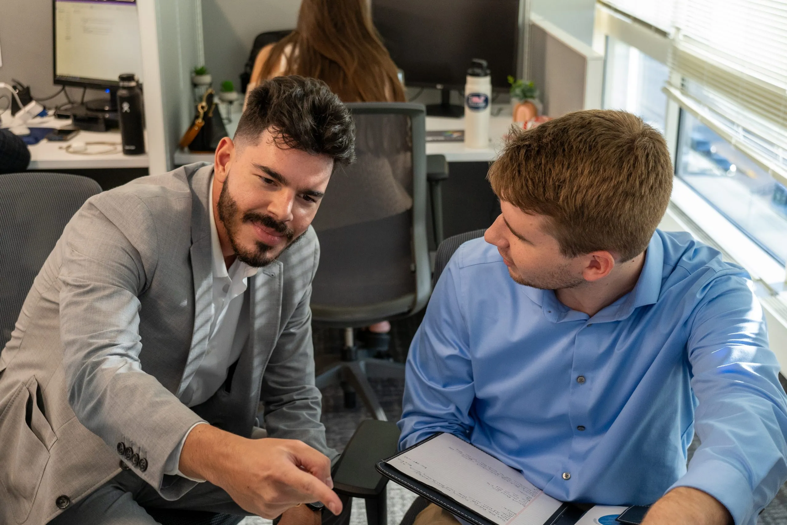 Two colleagues in business attire talk in an office, one pointing while the other holds a notepad